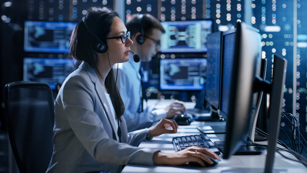 Female working in a Technical Support Team Gives Instructions with the Help of the Headsets. In the Background People Working and Monitors Show Various Information