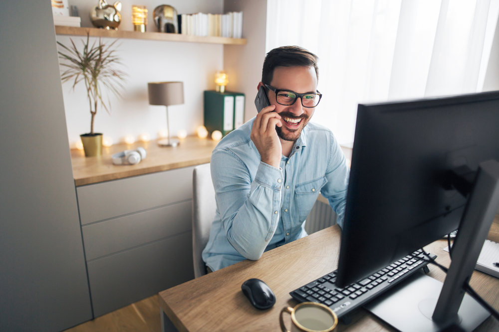 Smiling handsome freelancer working remotely from home. He is speaking on the phone.