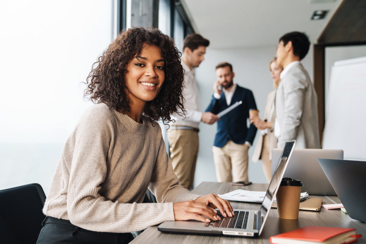 young confident businesswoman sitting at the office table with group of colleagues in the background, working on laptop computer